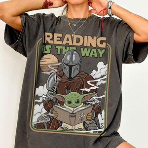 Puede incluir: Camiseta gris oscuro con un gr&aacute;fico de The Mandalorian y Grogu leyendo un libro, con el texto "READING IS THE WAY". El dise&ntilde;o incluye una puesta de sol y nubes, enmarcado en un borde verde y marr&oacute;n. La camiseta tiene mangas cortas.