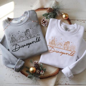 May include: Two sweatshirts, one gray and one white, each featuring a line drawing of the Disneyworld skyline. The word "Disneyworld" is written in a cursive font below the drawing. The gray sweatshirt has black text, while the white one has orange text.