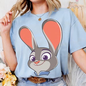 Puede incluir: Camiseta azul claro con un dibujo animado de un conejo gris sonriente. El conejo tiene orejas rosas y ojos morados. La camiseta de manga corta es de color liso.