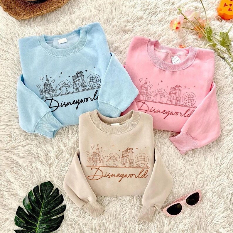 May include: Three crewneck sweatshirts in light blue, pink, and beige. Each features a black line drawing of a castle, tree, and globe, with the word "Disneyworld" in script below. The sweatshirts are displayed on a white, fluffy surface.