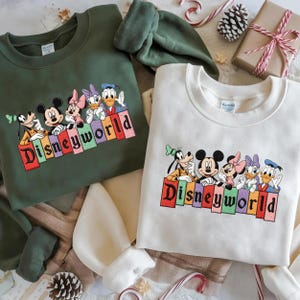 May include: Two crewneck sweatshirts, one forest green and one off-white, featuring a Disney World design. The design includes Mickey Mouse, Minnie Mouse, Donald Duck, and Goofy. The word "Disneyworld" is embroidered in colorful letters.