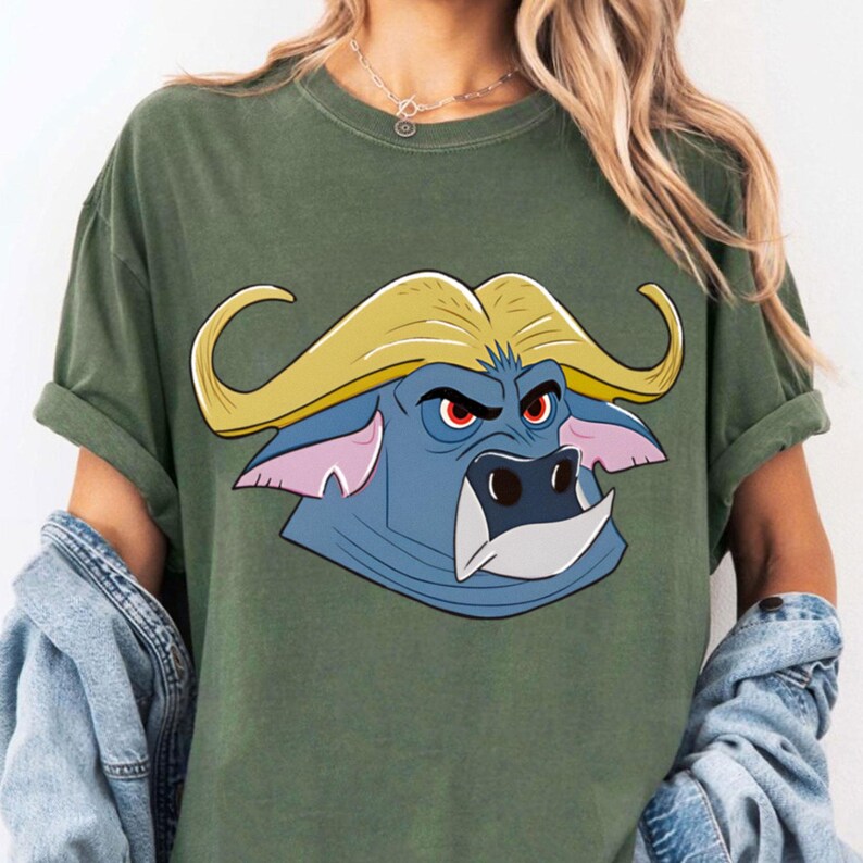 May include: Olive green t-shirt featuring a cartoon buffalo head with yellow horns, blue face, and red eyes. The buffalo has a stern expression. The shirt has short sleeves and a relaxed fit.
