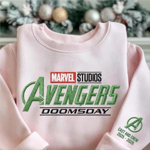May include: A light pink sweatshirt featuring the Marvel Studios Avengers logo in green and red embroidery. The word "DOOMSDAY" is embroidered below the logo. The sleeve cuff displays the Avengers "A" logo and the text "CAST AND CREW 2025-2026".