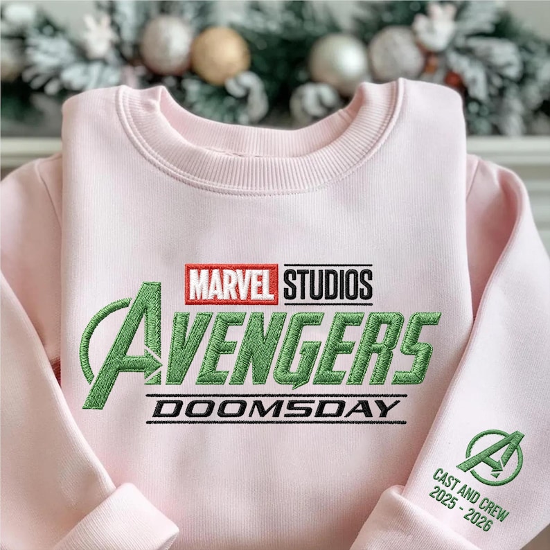 May include: A light pink sweatshirt featuring the Marvel Studios Avengers logo in green and red embroidery. The word "DOOMSDAY" is embroidered below the logo. The sleeve cuff displays the Avengers "A" logo and the text "CAST AND CREW 2025-2026".