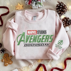 May include: A light pink sweatshirt featuring the Marvel Studios Avengers logo in green and black embroidery. The text "DOOMSDAY" is below the logo. The right sleeve has the Avengers "A" logo and "CAST AND CREW 2025-2026" in green.