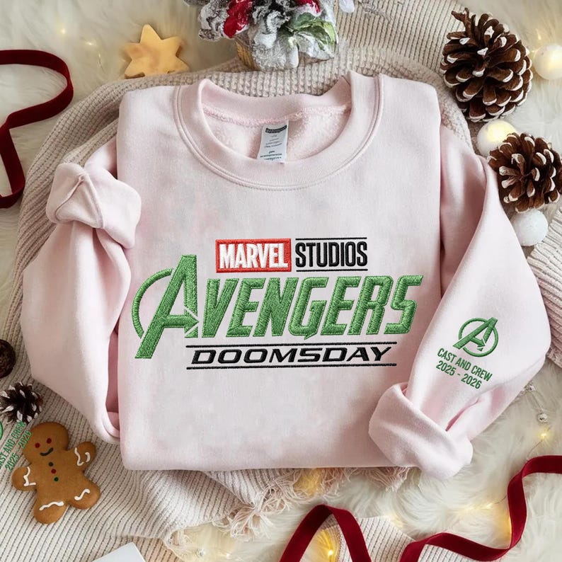 May include: A light pink sweatshirt featuring the Marvel Studios Avengers logo in green and black embroidery. The text "DOOMSDAY" is below the logo. The right sleeve has the Avengers "A" logo and "CAST AND CREW 2025-2026" in green.
