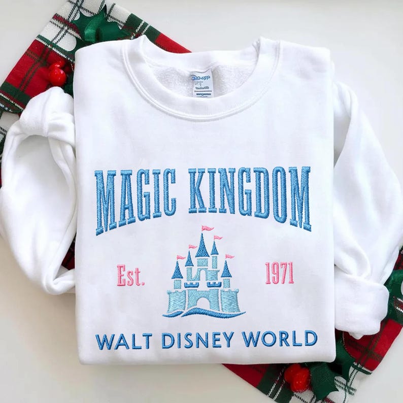 May include: A white sweatshirt with the words "MAGIC KINGDOM" in blue, arched above a light blue castle with pink flags. Below the castle is "WALT DISNEY WORLD". The year "1971" is in pink.