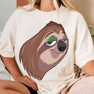 May include: Cream-colored t-shirt featuring a cartoon sloth face with brown fur, green eyes, and a black nose. The sloth has a relaxed expression. The shirt is made of a soft material.