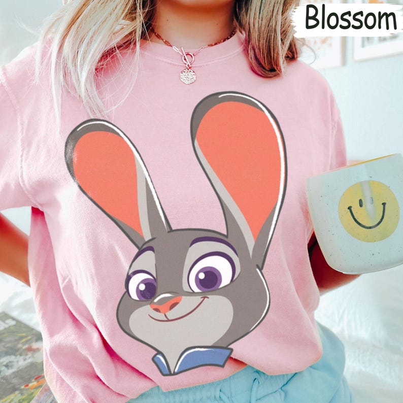 Puede incluir: Camiseta rosa claro con un dibujo animado de una cara de conejo. El conejo tiene orejas grandes, pelaje gris y un cuello azul. La palabra "Blossom" est&aacute; en la esquina superior derecha. Una taza blanca con una carita sonriente amarilla est&aacute; en la imagen.