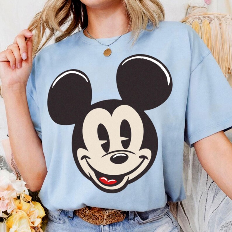 May include: A light blue t-shirt featuring a large graphic of Mickey Mouse's face. The design includes black ears, a smiling face with a beige muzzle, and a red mouth. The shirt has short sleeves and a crew neck.