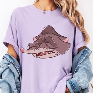 Peut inclure: T-shirt lilas avec un visage de possum de dessin anim&eacute;. Le possum a les yeux ferm&eacute;s et la bouche ouverte, dans les tons marron et rose. Le t-shirt a des manches retrouss&eacute;es.