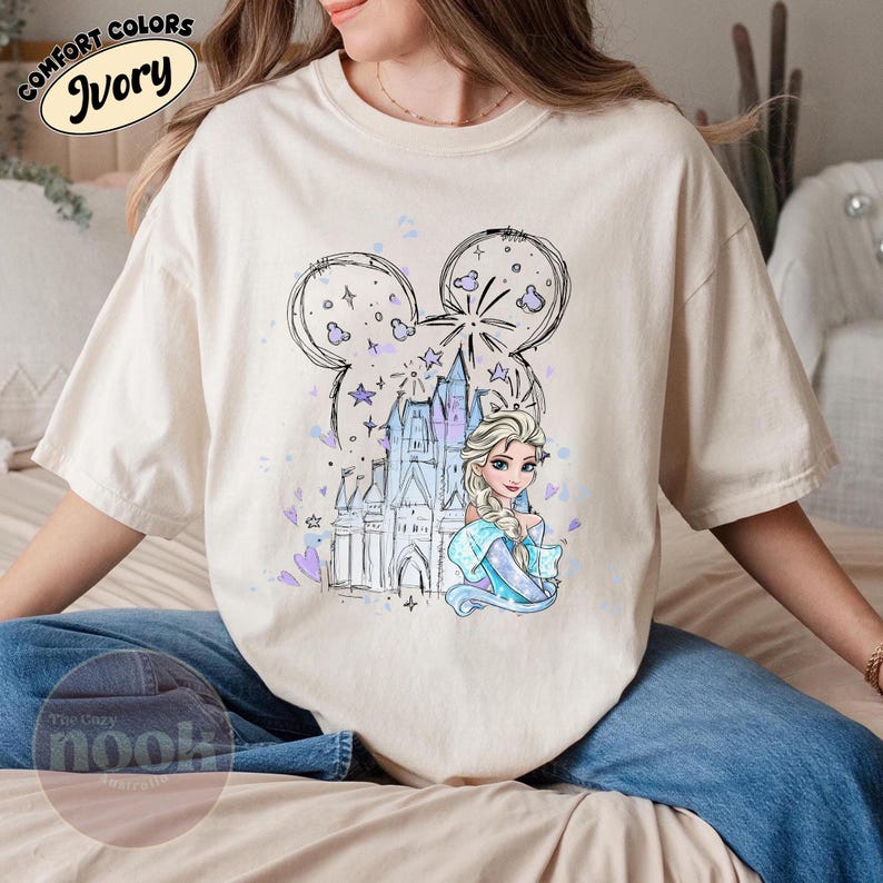 May include: Ivory-colored t-shirt featuring a whimsical design. The graphic includes a castle, fireworks, and a princess, all within a Mickey Mouse ear outline. The text "COMFORT COLORS Ivory" is printed at the top.