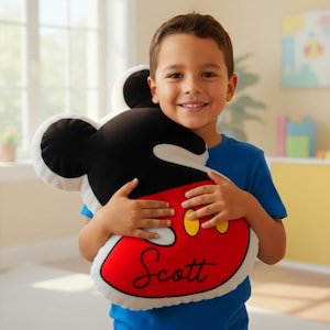 Op de afbeelding: Een Mickey Mouse-vormig kussen in zwart, wit, rood en geel. Het kussen heeft de naam "Scott" in sierlijke letters op het rode gedeelte. Een persoon met een blauwe top houdt het kussen vast. Een populair kinderartikel.