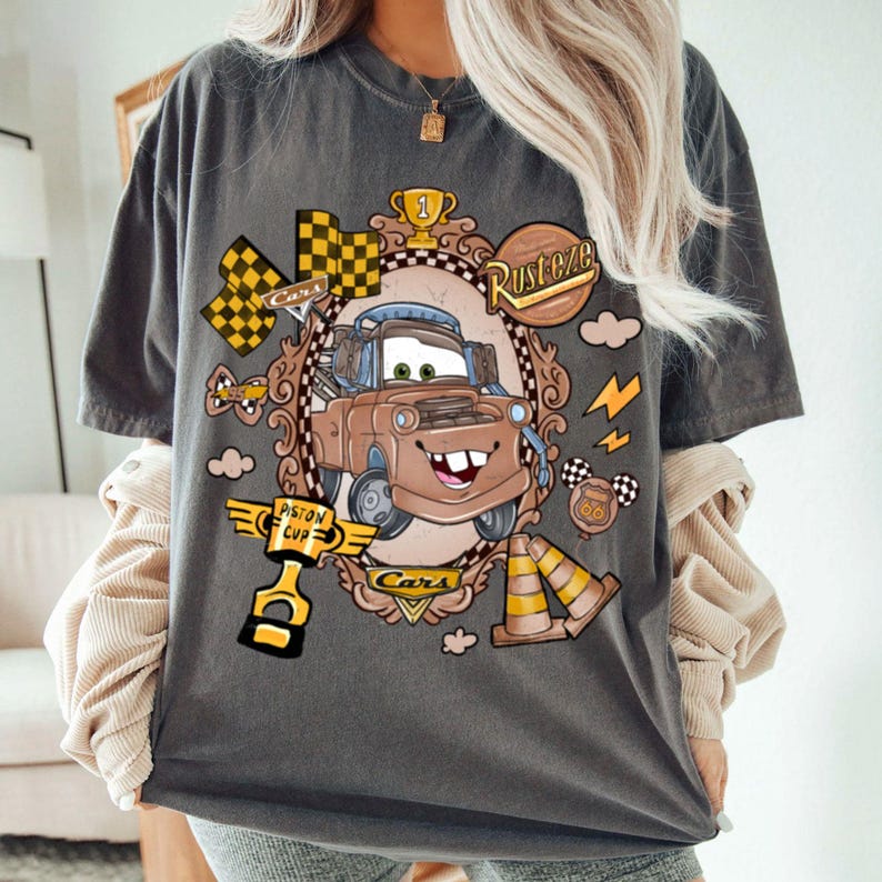 May include: Dark gray t-shirt featuring a cartoon image of Mater from the movie Cars. The graphic includes racing flags, lightning bolts, and the words "Rusteze" and "Piston Cup". The design is framed with a decorative border.