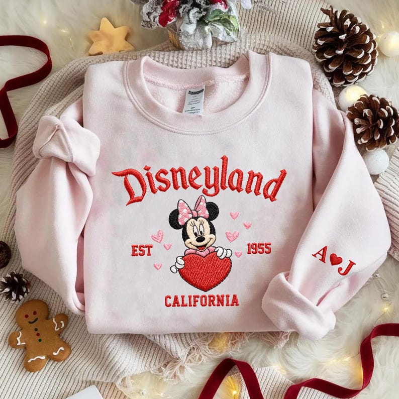 Puede incluir: Una sudadera rosa claro de Disneyland con texto bordado en rojo y un gr&aacute;fico de Minnie Mouse sosteniendo un coraz&oacute;n rojo. La manga tiene las iniciales "A ❤️ J". La sudadera est&aacute; rodeada de decoraciones navide&ntilde;as.