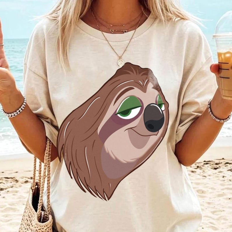May include: Beige t-shirt featuring a cartoon sloth with brown fur, green eyes, and a black nose. The sloth is smiling. The shirt is a casual style, suitable for everyday wear.