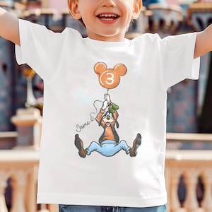 May include: White t-shirt featuring a cartoon of Goofy holding a balloon with the number 3. The balloon is orange and has a Mickey Mouse head shape. The name "James" is written in cursive below the cartoon.