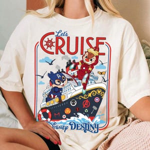 May include: Cream-colored t-shirt featuring a graphic of a cruise ship with cartoon characters dressed as superheroes. The shirt has the words "Let's Cruise" and "Disney Destiny" printed on it. The design includes red, blue, and yellow accents.