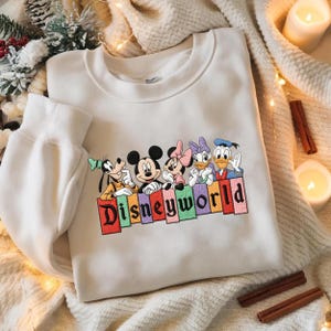 May include: A white sweatshirt featuring embroidered Disney characters: Goofy, Mickey Mouse, Minnie Mouse, Daisy Duck, and Donald Duck. The word "Disneyworld" is embroidered in colorful letters below the characters. The sweatshirt is on a cream-colored blanket.