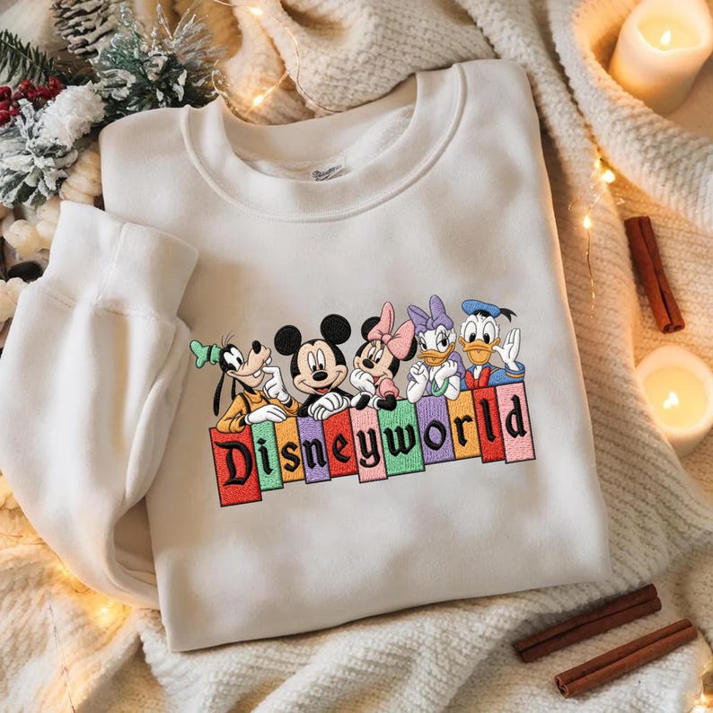 May include: A white sweatshirt featuring embroidered Disney characters: Goofy, Mickey Mouse, Minnie Mouse, Daisy Duck, and Donald Duck. The word "Disneyworld" is embroidered in colorful letters below the characters. The sweatshirt is on a cream-colored blanket.
