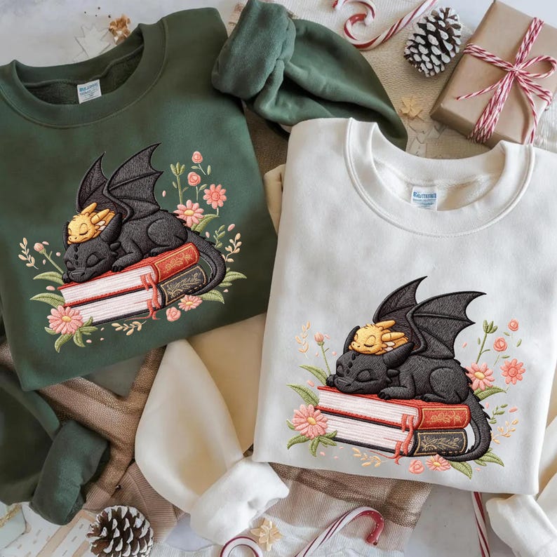 May include: Two sweatshirts, one green and one cream, each featuring an embroidered design of a sleeping dragon on a stack of books, surrounded by flowers. The dragon is black with a yellow head. The sweatshirts are a cozy, casual style.