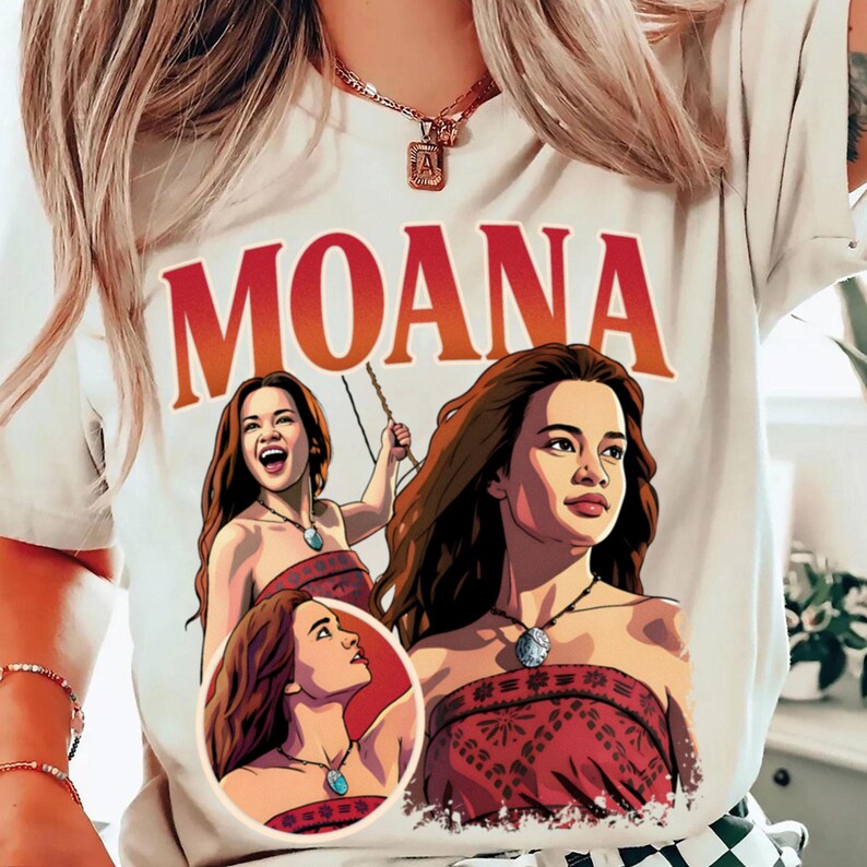 以下が含まれることがあります： 上部に大きな赤い文字でMOANAと書かれたクリーム色のTシャツ。テキストの下には、それぞれ柄入りの赤いトップスと青いペンダントネックレスを身に着けた、モアナというキャラクターの3つの異なるイラストが描かれています。