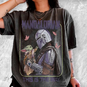May include: Dark gray t-shirt featuring a graphic of The Mandalorian and Grogu, with the text "MANDALORIAN" and "THIS IS THE WAY". The design includes a helmeted figure and a small green creature, with pink butterflies.