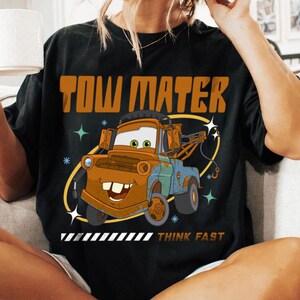 May include: A black t-shirt featuring a graphic of the character Tow Mater from the movie Cars. The design includes the text TOW MATER in large orange letters above the truck and the text THINK FAST in smaller letters below the truck graphic.