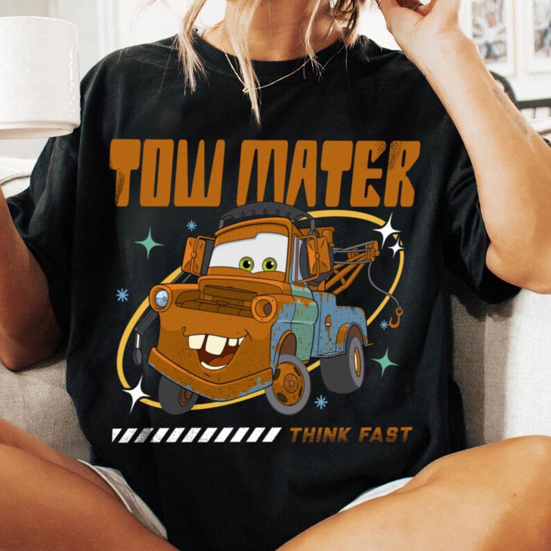 May include: A black t-shirt featuring a graphic of the character Tow Mater from the movie Cars. The design includes the text TOW MATER in large orange letters above the truck and the text THINK FAST in smaller letters below the truck graphic.
