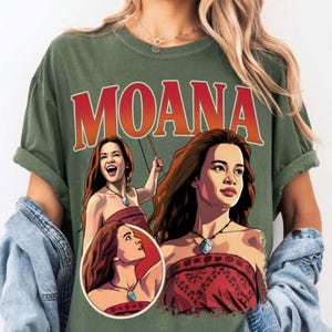 以下が含まれることがあります： オリーブグリーンのTシャツで、キャラクター「モアナ」の大きなグラフィックプリントが特徴です。デザインには太い赤文字で「MOANA」と書かれており、赤い柄のトップスと青いネックレスを身に着けたキャラクターの3つの異なるイラストが描かれています。