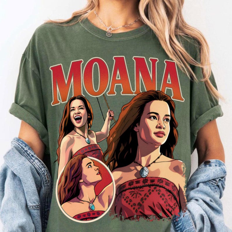 以下が含まれることがあります： オリーブグリーンのTシャツで、キャラクター「モアナ」の大きなグラフィックプリントが特徴です。デザインには太い赤文字で「MOANA」と書かれており、赤い柄のトップスと青いネックレスを身に着けたキャラクターの3つの異なるイラストが描かれています。