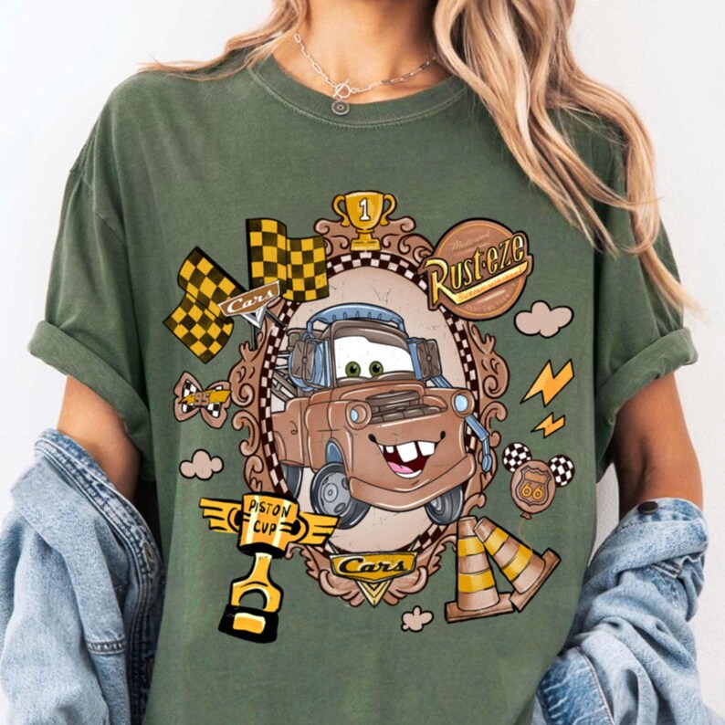 May include: Green t-shirt featuring a cartoon image of Mater from the movie Cars. The design includes racing flags, a trophy, and the words "Rusteze" and "Piston Cup". The shirt has short sleeves.