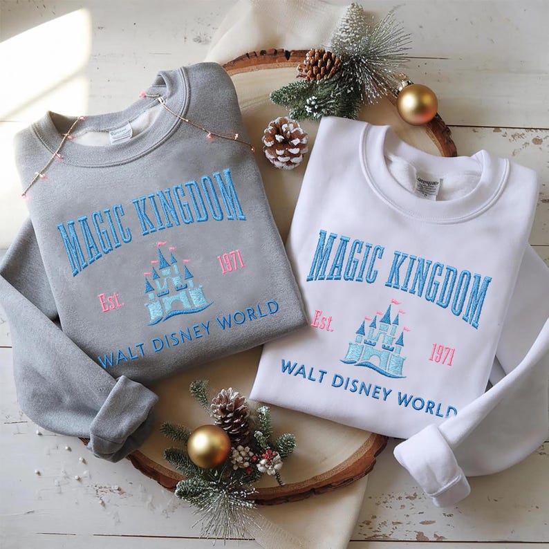 May include: Two sweatshirts, one gray and one white, with the words "MAGIC KINGDOM" embroidered in blue. Below the text is a castle design and the words "WALT DISNEY WORLD" and "Est. 1971" in pink. The sweatshirts are on a wooden surface.