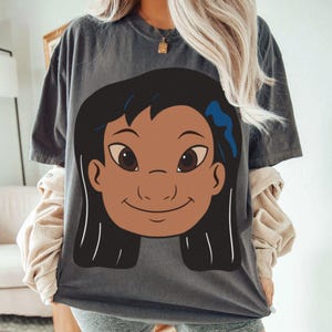 Puede incluir: Camiseta gris oscuro con una ilustraci&oacute;n de dibujos animados de la cara de una ni&ntilde;a con cabello negro, piel morena y un lazo azul. El gr&aacute;fico est&aacute; centrado en la parte delantera de la camiseta. La camiseta tiene mangas cortas y cuello redondo.
