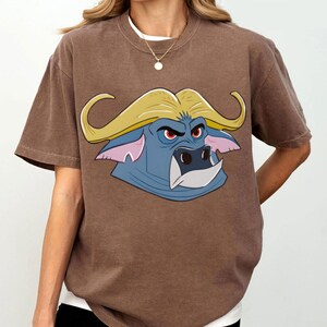 May include: Brown t-shirt featuring a cartoon buffalo head with a blue face, yellow hair, and red eyes. The buffalo graphic is centered on the shirt. The shirt has short sleeves and a crew neck.