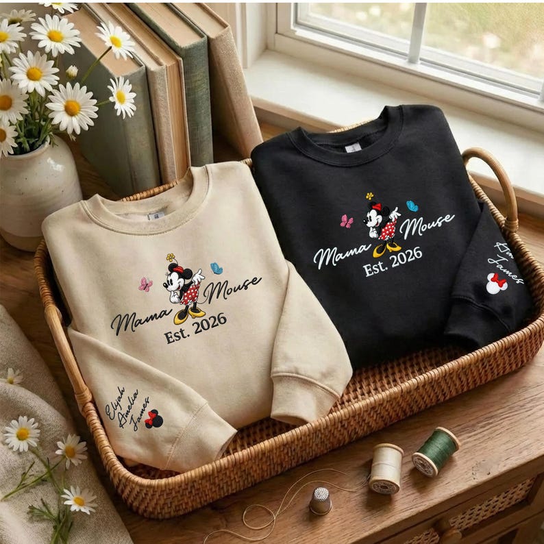 K&ouml;nnte beinhalten: Zwei Sweatshirts, eines beige und eines schwarzen, mit einem Minnie Mouse-Design und dem Text "Mama Mouse Est. 2026". Das beige Sweatshirt hat "Elijah Amelia James" auf dem &Auml;rmel. Sie liegen in einem Weidenkorb.