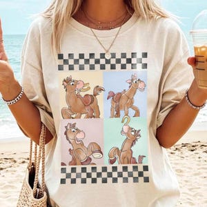 May include: Beige t-shirt featuring a four-panel design of a cartoon horse in various poses. The design is framed by a black and white checkered pattern. The horse is brown with a saddle and a horseshoe. The shirt is a casual style.