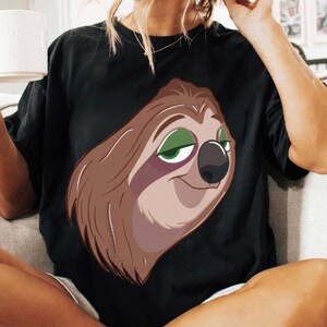 May include: Black t-shirt featuring a cartoon sloth with brown fur, green eyes, and a black nose. The sloth is smiling. The shirt is a casual style.