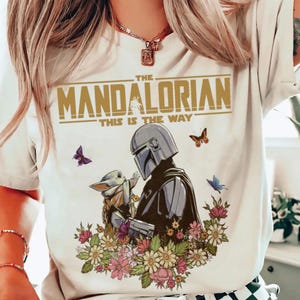 May include: White t-shirt featuring a graphic of The Mandalorian and Grogu surrounded by flowers and butterflies. The text "THE MANDALORIAN" and "THIS IS THE WAY" are printed in gold.