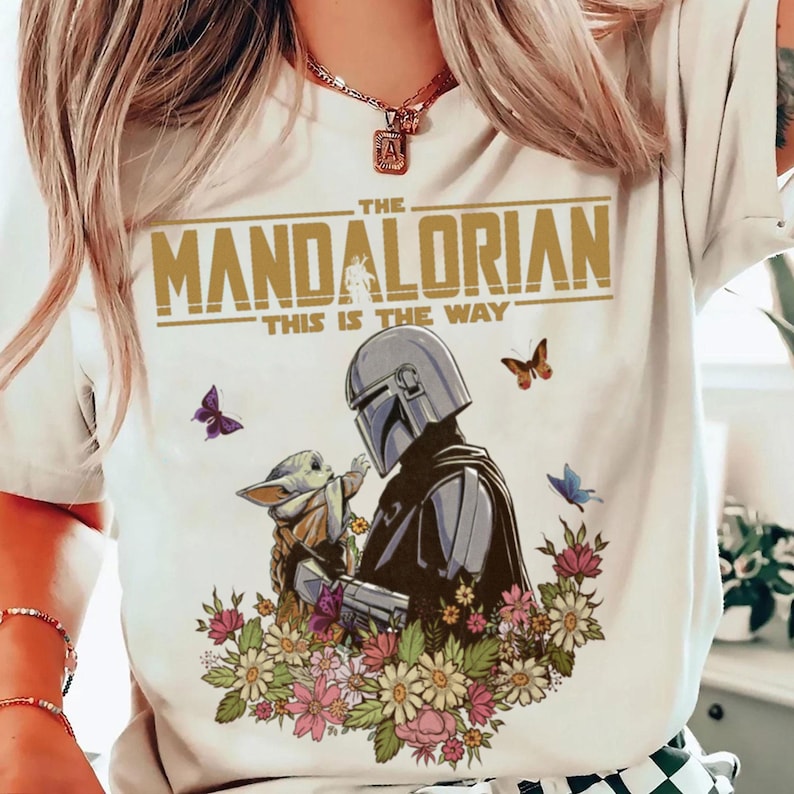 May include: White t-shirt featuring a graphic of The Mandalorian and Grogu surrounded by flowers and butterflies. The text "THE MANDALORIAN" and "THIS IS THE WAY" are printed in gold.
