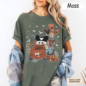 May include: Moss green t-shirt featuring a cartoon tow truck with Mickey Mouse ears and Halloween-themed balloons, pumpkins, and ghosts. The shirt has a distressed look and the text "The Cozy Nook" is visible. The word "Moss" is printed on the top right.