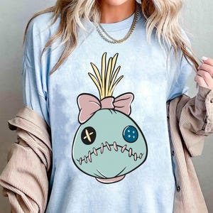 May include: Light blue t-shirt featuring a cartoon doll face design. The doll face is light blue with a pink bow, yellow hair, and stitched details. The design includes a black cross, a blue button, and a stitched mouth.