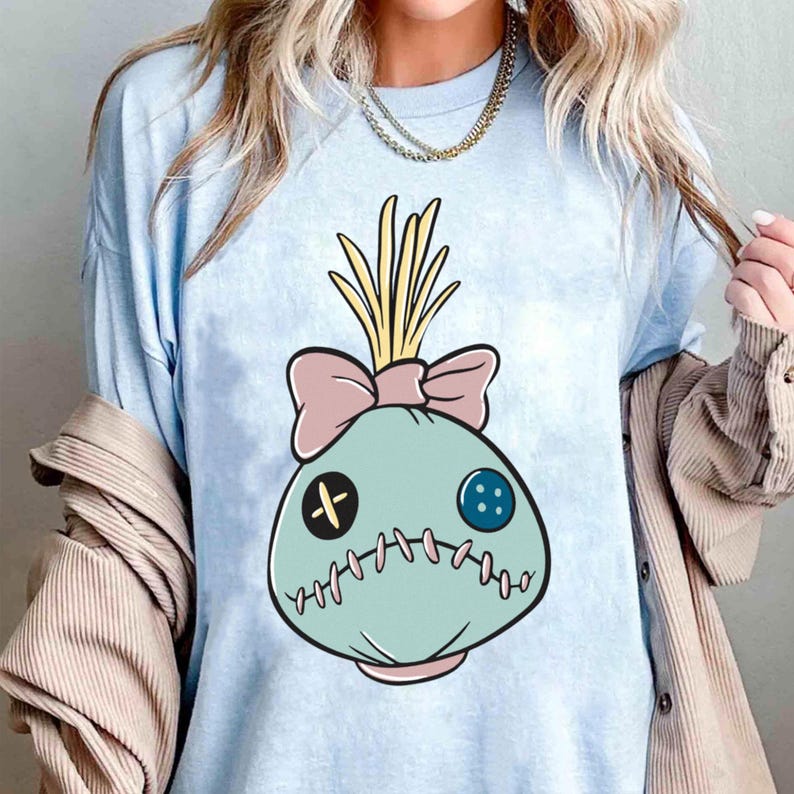 May include: Light blue t-shirt featuring a cartoon doll face design. The doll face is light blue with a pink bow, yellow hair, and stitched details. The design includes a black cross, a blue button, and a stitched mouth.