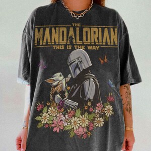 May include: Dark gray t-shirt featuring a graphic of The Mandalorian and Grogu surrounded by flowers and butterflies. The text "THE MANDALORIAN" and "THIS IS THE WAY" are printed across the top of the shirt in gold.