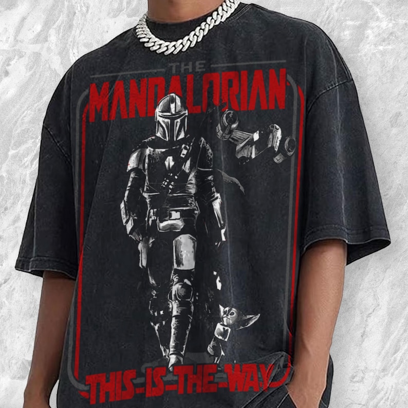 May include: Black t-shirt featuring a graphic of The Mandalorian with the text "THE MANDALORIAN" and "THIS IS THE WAY" in red. The design includes an illustration of the Mandalorian, a ship, and Grogu. The shirt has a vintage, distressed look.