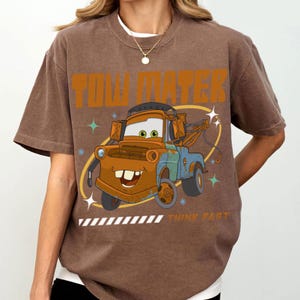 May include: A brown short-sleeve t-shirt featuring a graphic of the character Tow Mater from the movie Cars. The design includes the text TOW MATER at the top and THINK FAST at the bottom, surrounded by star accents and a yellow oval outline.