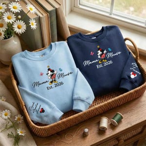K&ouml;nnte beinhalten: Zwei Sweatshirts in einem Weidenkorb. Eines ist hellblau, das andere marineblau. Beide haben eine Minnie Mouse-Grafik mit dem Text "Mama Mouse Est. 2026." Das hellblaue Sweatshirt hat die Namen "Elyah Amelia James" auf dem &Auml;rmel.