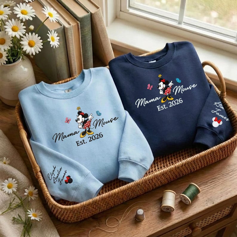K&ouml;nnte beinhalten: Zwei Sweatshirts in einem Weidenkorb. Eines ist hellblau, das andere marineblau. Beide haben eine Minnie Mouse-Grafik mit dem Text "Mama Mouse Est. 2026." Das hellblaue Sweatshirt hat die Namen "Elyah Amelia James" auf dem &Auml;rmel.