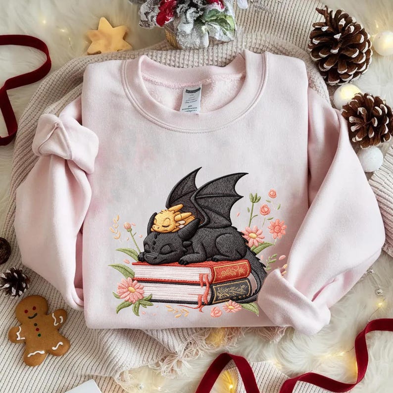 May include: A light pink sweatshirt featuring an embroidered design of a black dragon and a yellow dragon sleeping on a stack of books, surrounded by pink flowers. The sweatshirt has long sleeves and a crew neck.
