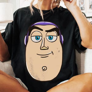 May include: Black t-shirt featuring a large cartoon graphic of Buzz Lightyear's face. The graphic includes a tan face, blue eyes, purple accents, and a smiling expression. The shirt is made of a soft material.